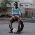 A young boy sits on a tire with a soccer ball in a quiet street, evoking a sense of play and solitude.