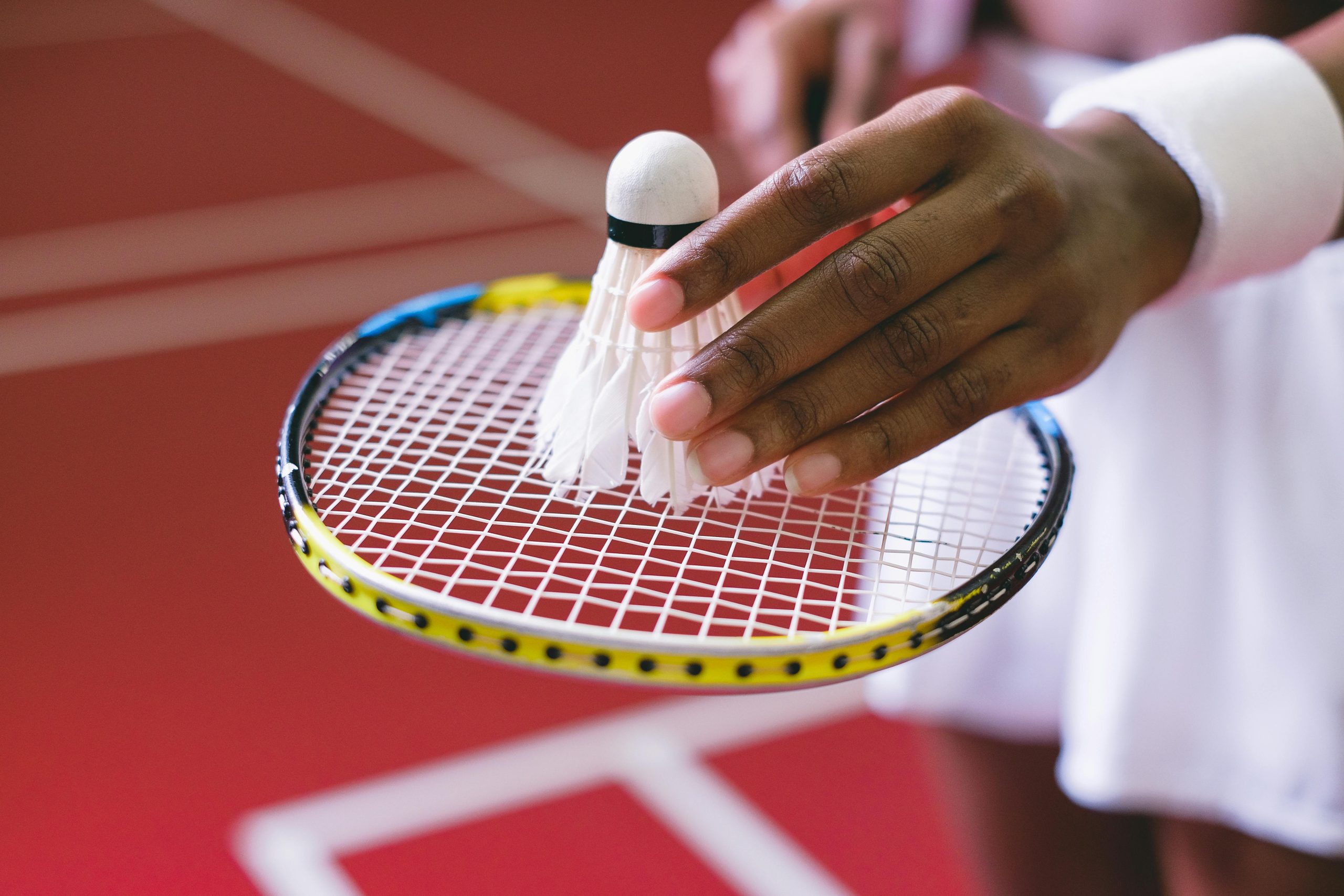 A detailed view of a hand holding a shuttlecock over a badminton racket, on an indoor court.