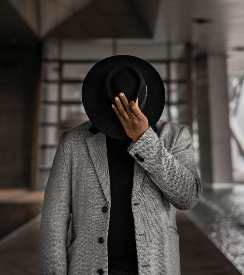 Elegant photo of a man in a formal coat and fedora hat posing indoors.